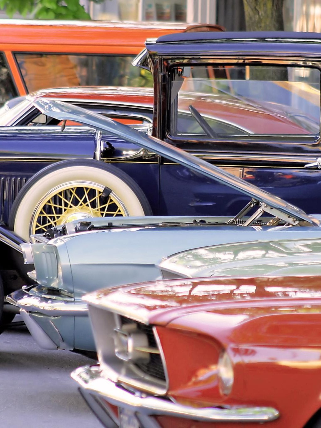 Eine Reihe von Oldtimern, darunter ein orange-rotes Cabrio, ein grünes Auto mit offenem Motor und ein blaues Auto mit Weißwandreifen.