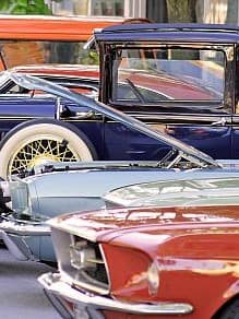 Eine Reihe von Oldtimern, darunter ein orange-rotes Cabrio, ein grünes Auto mit offenem Motor und ein blaues Auto mit Weißwandreifen.