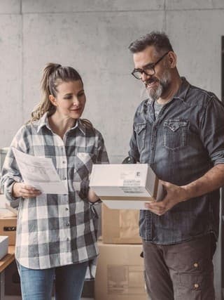 Eine Frau und ein Mann stehen in einem Lager voller Pakete. Die Frau hält ein Dokument in der Hand, während der Mann ein Paket hält. Beide blicken lächelnd auf das Paket.
