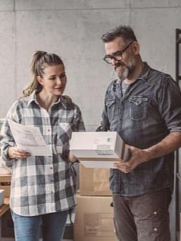 Eine Frau und ein Mann stehen in einem Lager voller Pakete. Die Frau hält ein Dokument in der Hand, während der Mann ein Paket hält. Beide blicken lächelnd auf das Paket.
