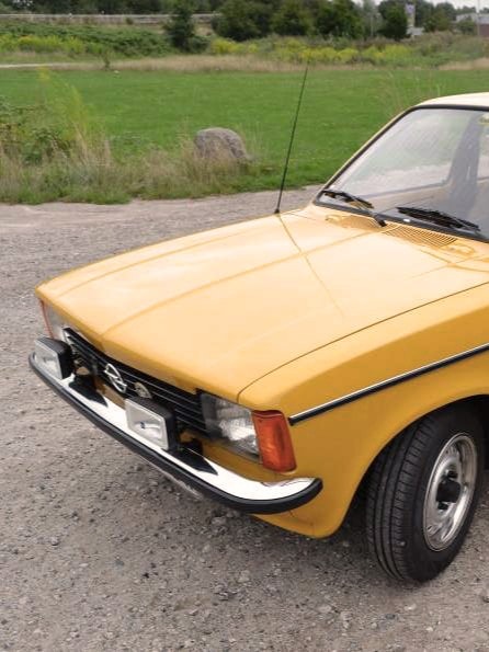Ein gelbes Vintage-Auto, vermutlich ein Opel Kadett aus den 1970er Jahren, steht auf einem Kiesweg mit einer Wiese im Hintergrund.