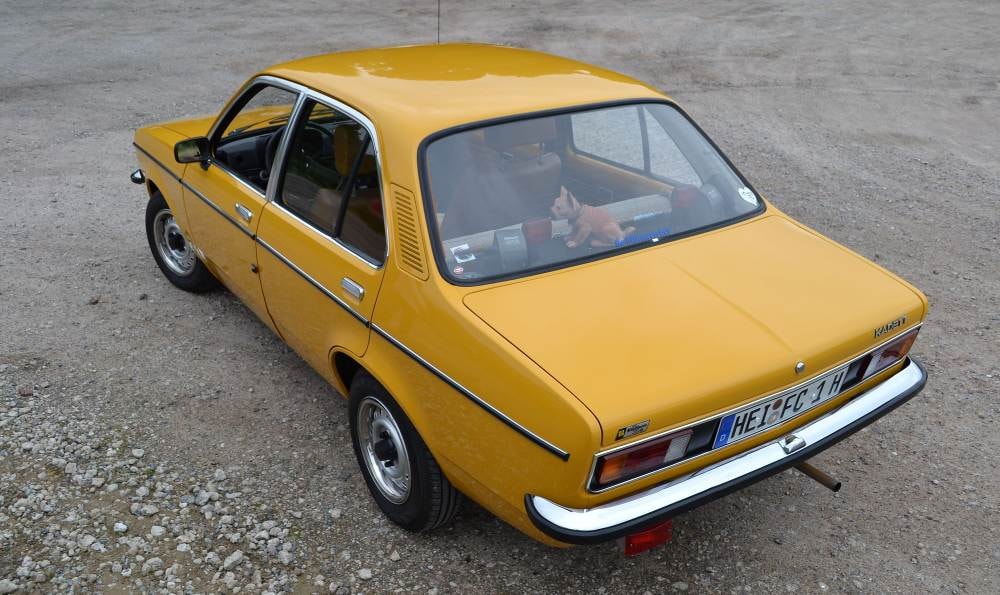 Gelbes Vintage-Auto mit kantigem Design und schwarzen Zierleisten, geparkt auf einem Kiesplatz.