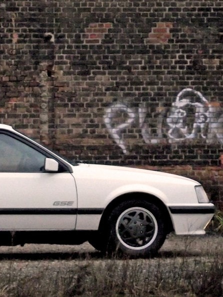 Weißer Opel Monza GSE parkt vor einer Backsteinwand mit Graffiti von Jens Tanz.