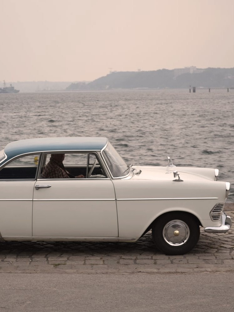Weißes klassisches Auto mit blauem Dach und Chromdetails, geparkt auf Kopfsteinpflaster nahe einem Gewässer.