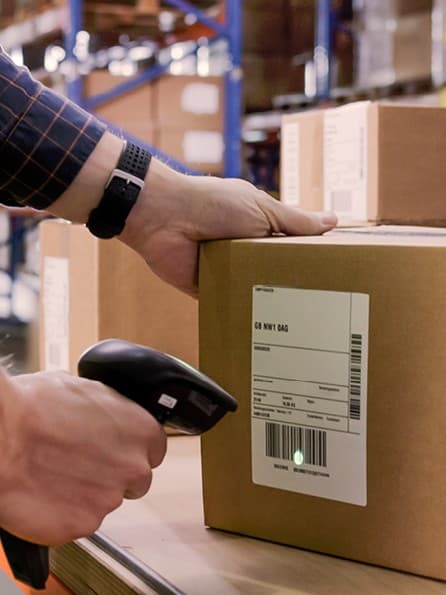 Paket liegt auf einem Tisch in einem Warenlager. Ein Mann in dunkelblauem Hemd hält einen Scanner in der rechten Hand und scannt das Paket en.