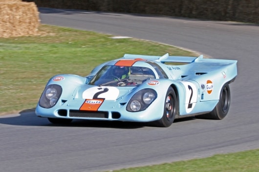 Ein hellblauer Oldtimer-Rennwagen Porsche 917k mit einem Gulf-Logo, der Startnummer 2 und eleganten Kurven rast über eine Rennstrecke, die von grünem Gras und einer Kurve aus Heuballen umgeben ist.