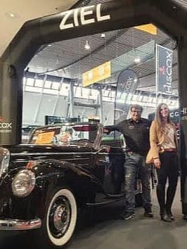 Klassischer schwarzer Wagen auf einer Messe unter einem ZIEL-Bogen, umgeben von vier Personen; HISCOX-Banner im Hintergrund.