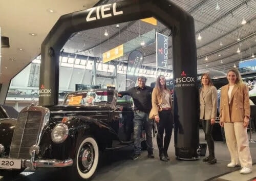 Klassischer schwarzer Wagen auf einer Messe unter einem ZIEL-Bogen, umgeben von vier Personen; HISCOX-Banner im Hintergrund.
