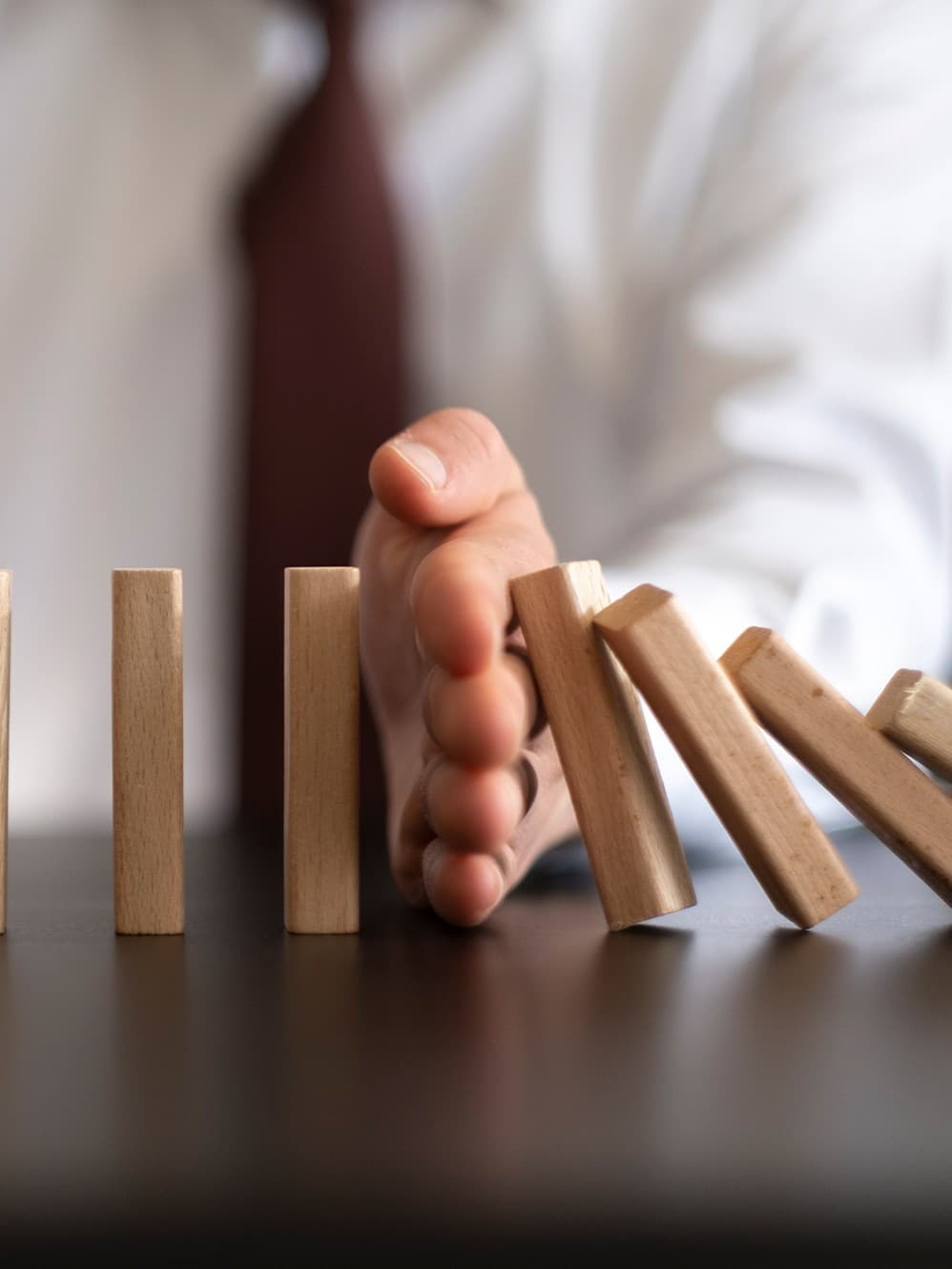 Zwei Hände halten fallende Dominosteine mitten in ihrer Bewegung auf einem dunklen Tisch an und symbolisieren so Eingreifen und Kontrolle. Die Szene vermittelt ein Gefühl der Ruhe und Prävention.