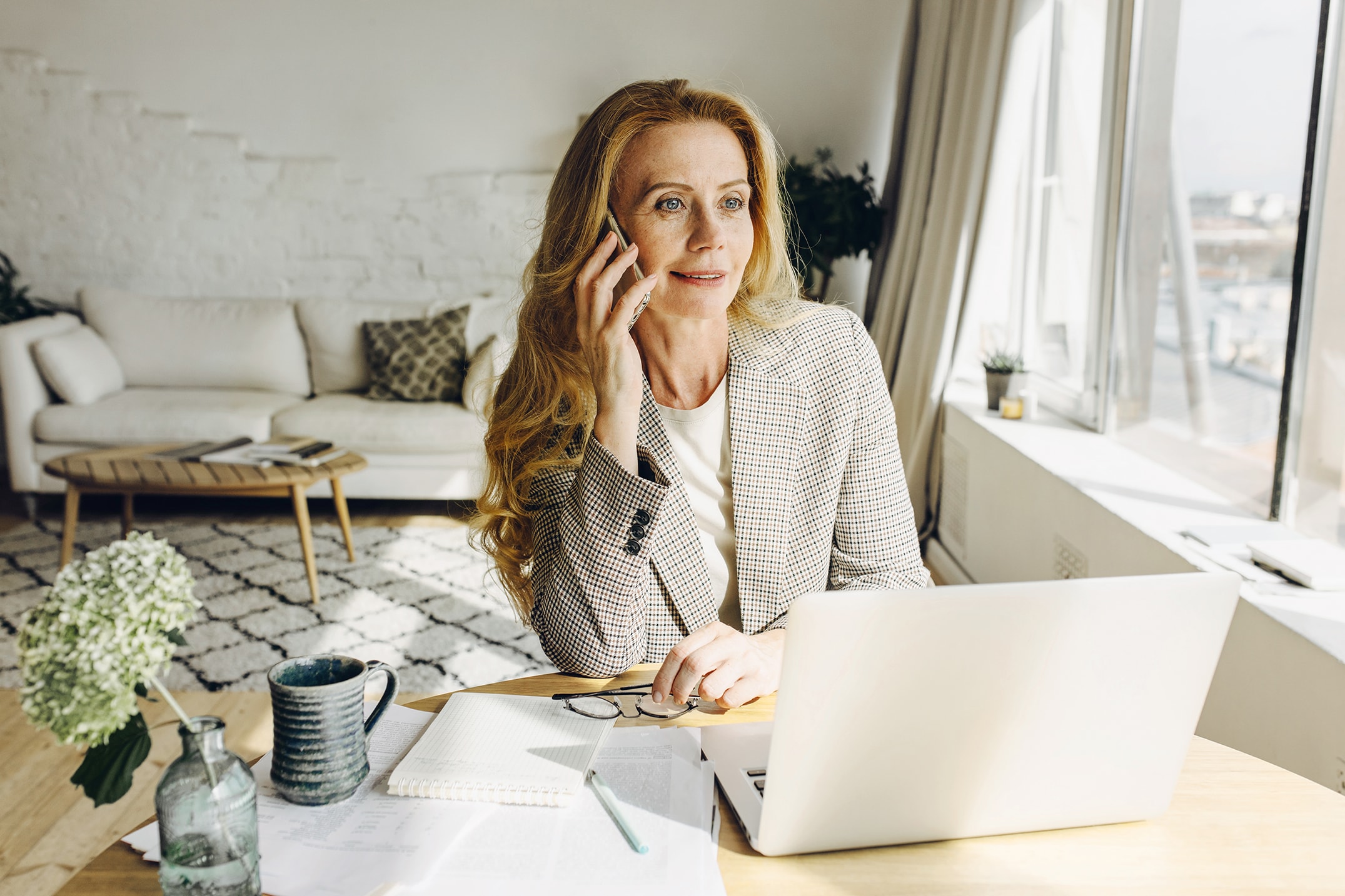Eine Frau mit langen roten Haaren sitzt an einem Schreibtisch und lächelt, während sie telefoniert. Sie befindet sich in einem hellen, modernen Wohnzimmer und wirkt konzentriert und zufrieden.