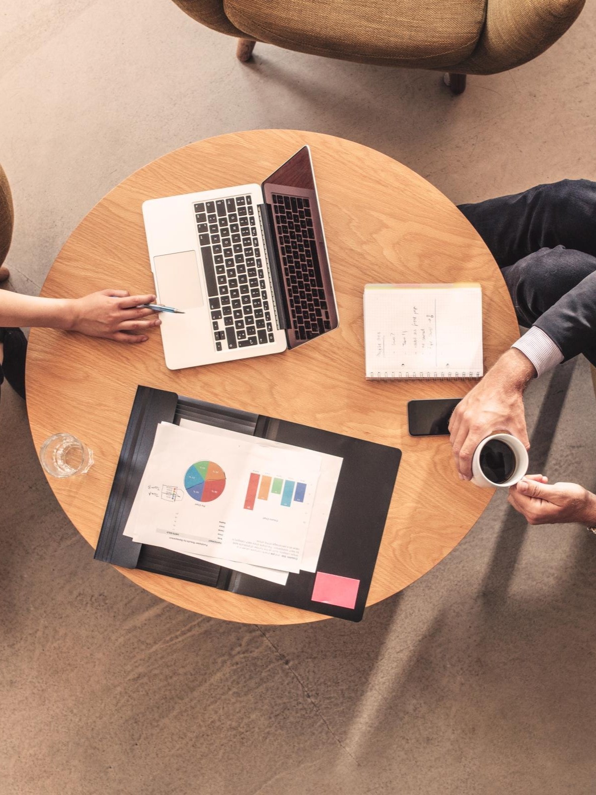 Zwei Personen sitzen an einem runden Holztisch. Auf dem Tisch liegen ein offener Laptop, ein Notizbuch mit handschriftlichen Notizen und eine Mappe mit Diagrammen und Grafiken. Eine Person zeigt mit einem Stift auf den Laptopbildschirm, die andere hält eine Tasse Kaffee.