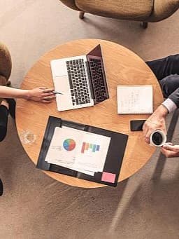 Zwei Personen sitzen an einem runden Holztisch. Auf dem Tisch liegen ein offener Laptop, ein Notizbuch mit handschriftlichen Notizen und eine Mappe mit Diagrammen und Grafiken. Eine Person zeigt mit einem Stift auf den Laptopbildschirm, die andere hält eine Tasse Kaffee.