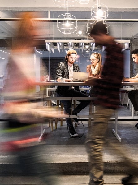 Ein volles Glasbüro eines Start-ups voll mit Mitarbeitern an ihren Laptops. Mitarbeiter laufen vorbei und gehen zu einem Meeting