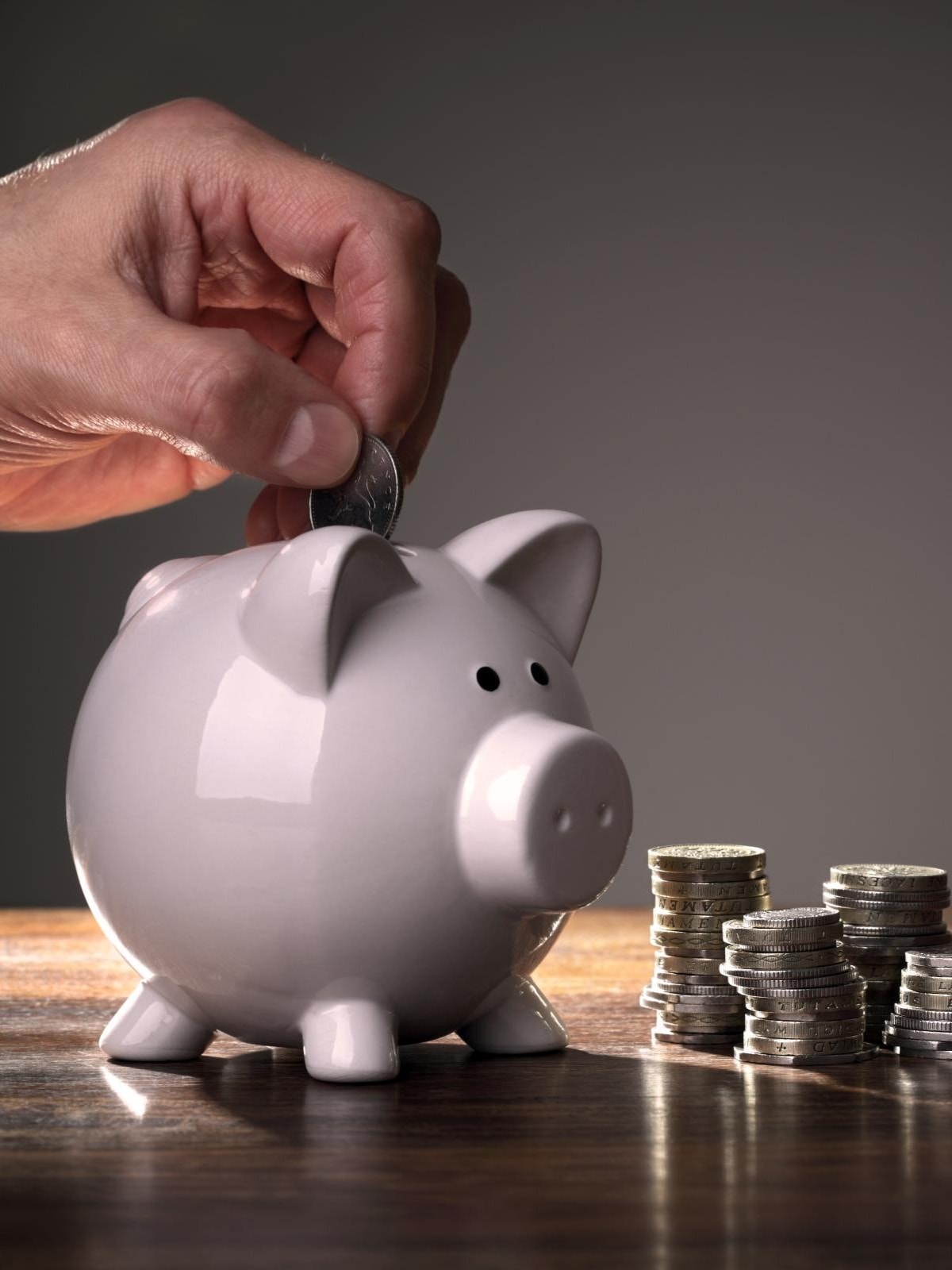 Eine Hand wirft eine Münze in ein weißes Sparschwein. Daneben stehen mehrere Münzstapel auf einer Holzoberfläche.