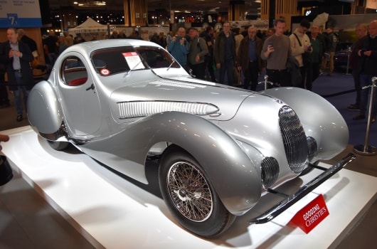 Ein eleganter silberner Oldtimer, der auf einer Plattform auf einer Automobilausstellung ausgestellt ist und von einer Menschenmenge umringt wird. Die Rundungen und Details des Wagens strahlen Eleganz und Nostalgie aus.