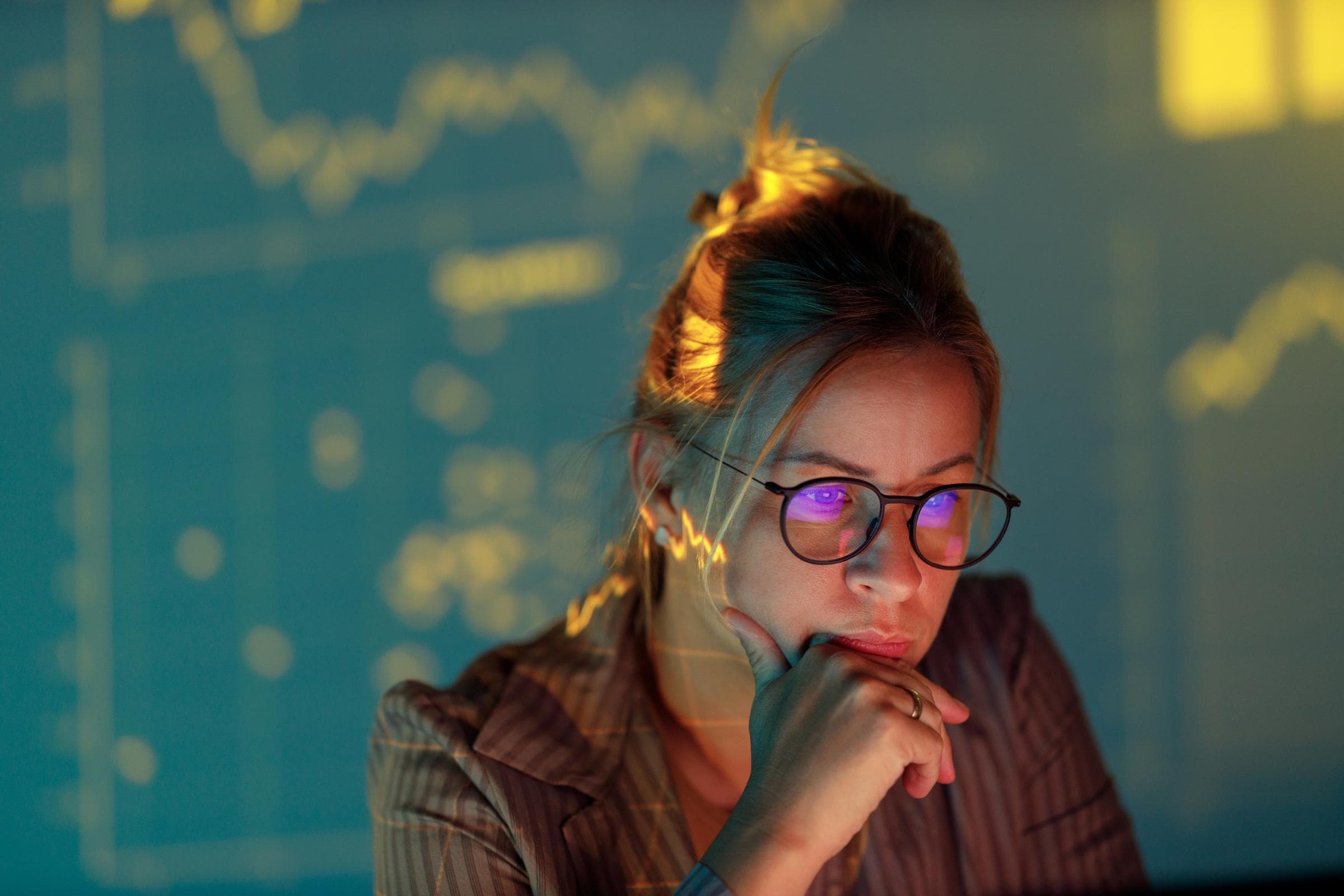 Eine Frau mit Brille blickt konzentriert auf einen Bildschirm, der von Finanzdiagrammen beleuchtet wird, die im Hintergrund projiziert werden. Die Szene strahlt eine nachdenkliche, analytische Atmosphäre aus.