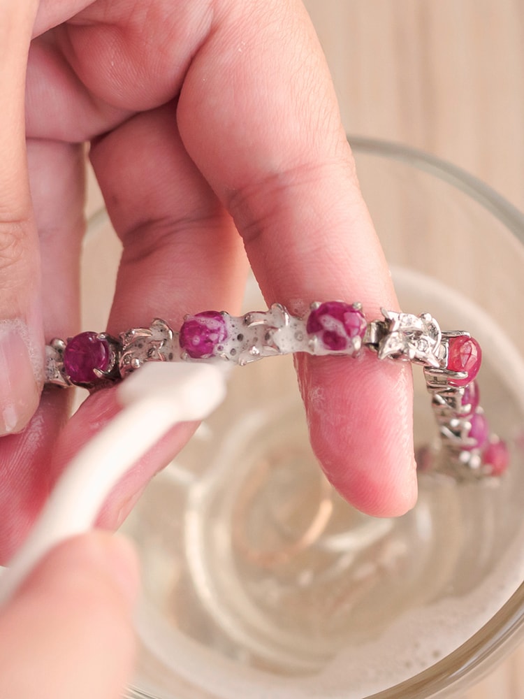 Ein Armband mit Edelsteinen wird mit einer Bürste über einem Glas Wasser gereinigt