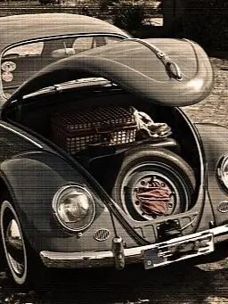 Ein klassischer VW Käfer mit geöffneter vorderer Haube, die einen Ersatzreifen, ein rotes Tuch und einen Picknickkorb im Kofferraum zeigt. Das Auto steht auf einem gepflasterten Weg, umgeben von Blumen.