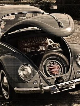 Ein klassischer VW Käfer mit geöffneter vorderer Haube, die einen Ersatzreifen, ein rotes Tuch und einen Picknickkorb im Kofferraum zeigt. Das Auto steht auf einem gepflasterten Weg, umgeben von Blumen.