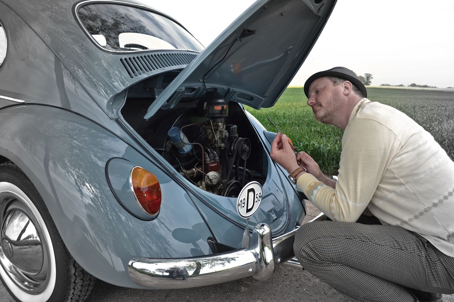 Eine Person arbeitet am Motor eines klassischen VW Käfers mit geöffneter Heckklappe. Die Person kniet neben dem Auto und benutzt Werkzeuge.