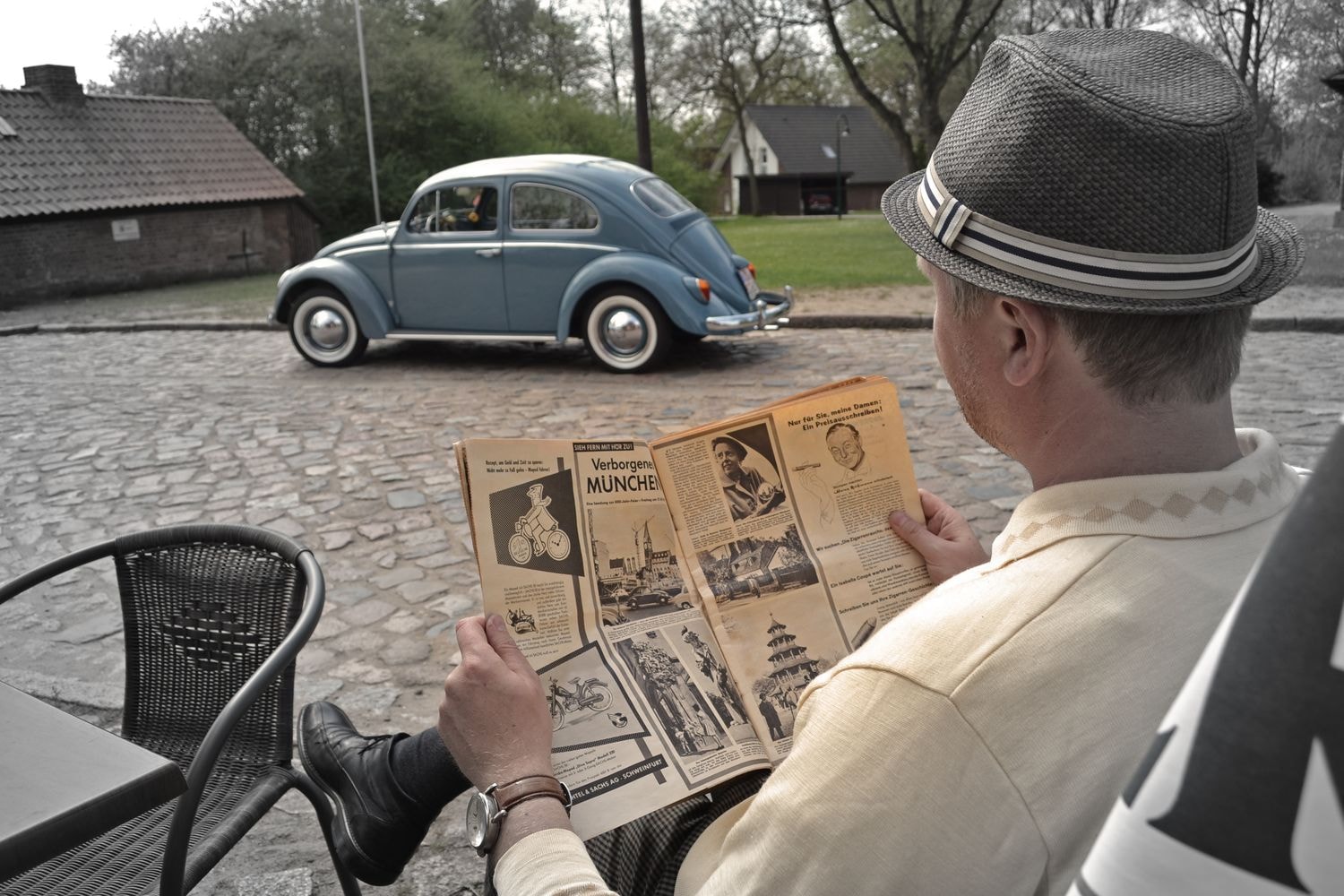 Eine Person sitzt in einem Straßencafé und liest eine Zeitung. Im Hintergrund steht ein blauer VW Käfer auf einem Kopfsteinpflaster. Bäume und Gebäude umrahmen die Szene.