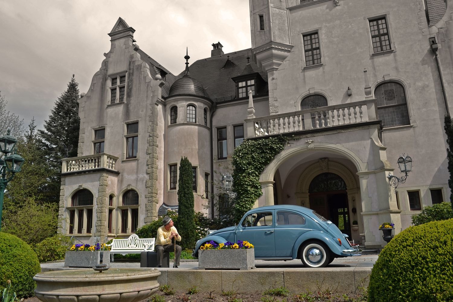 Ein historisches Gebäude mit Steinmauern, Rundbogenfenstern und einem Turm. Davor steht ein blauer Oldtimer, eine weiße Bank und eine Person mit Hut in heller Kleidung.