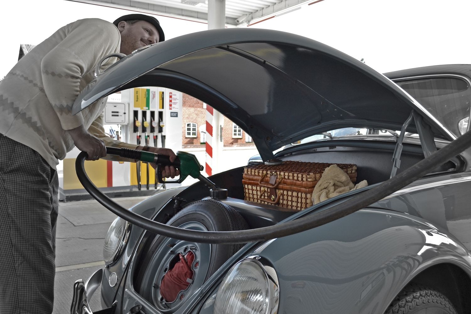 Eine Person tankt einen Oldtimer an einer Tankstelle. Die Motorhaube ist geöffnet und zeigt den Motor sowie einen Weidenkorb. Im Hintergrund sind mehrere Zapfsäulen zu sehen.