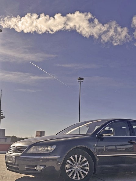 Eine schwarze VW Phaeton D1 3.0 TDI Individual 2007 parkt unter einem klaren blauen Himmel mit einer hellen Sonne in der Ecke. Im Hintergrund steigt Rauch aus hohen Industrieschornsteinen auf.