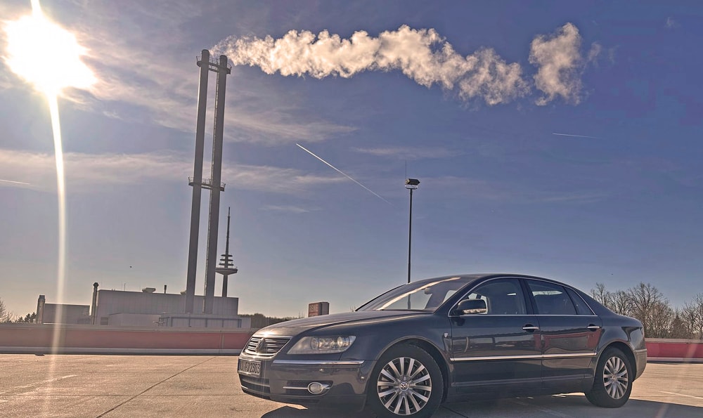 Eine schwarze VW Phaeton D1 3.0 TDI Individual 2007 parkt unter einem klaren blauen Himmel mit einer hellen Sonne in der Ecke. Im Hintergrund steigt Rauch aus hohen Industrieschornsteinen auf.