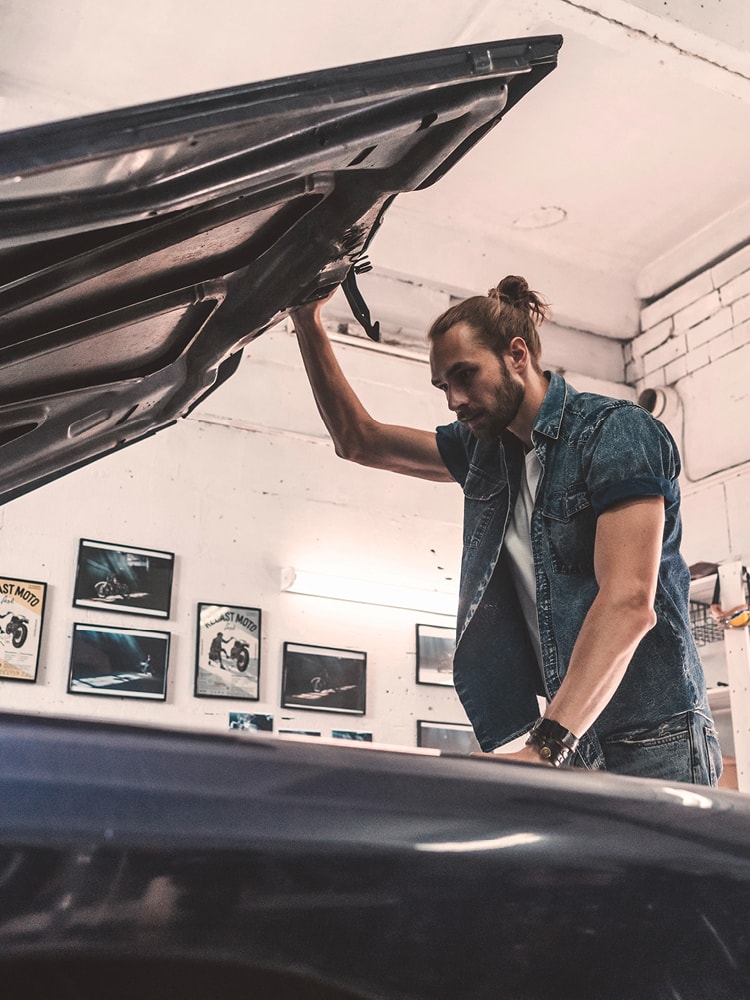 Ein Mann in Jeansweste steht in einer Garage und öffnet die Motorhaube seines Fahrzeugs