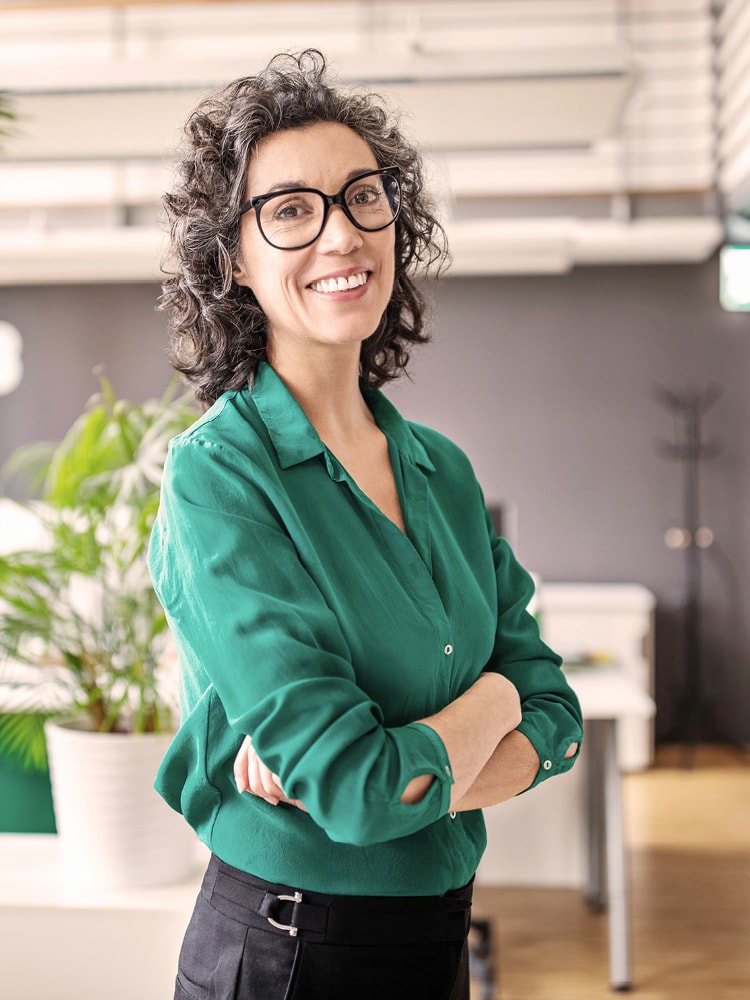 Eine Frau mit Locken und einer grünen Bluse steht in einem Büro, hat die Arme verschränkt und blickt lachend in die Kamera