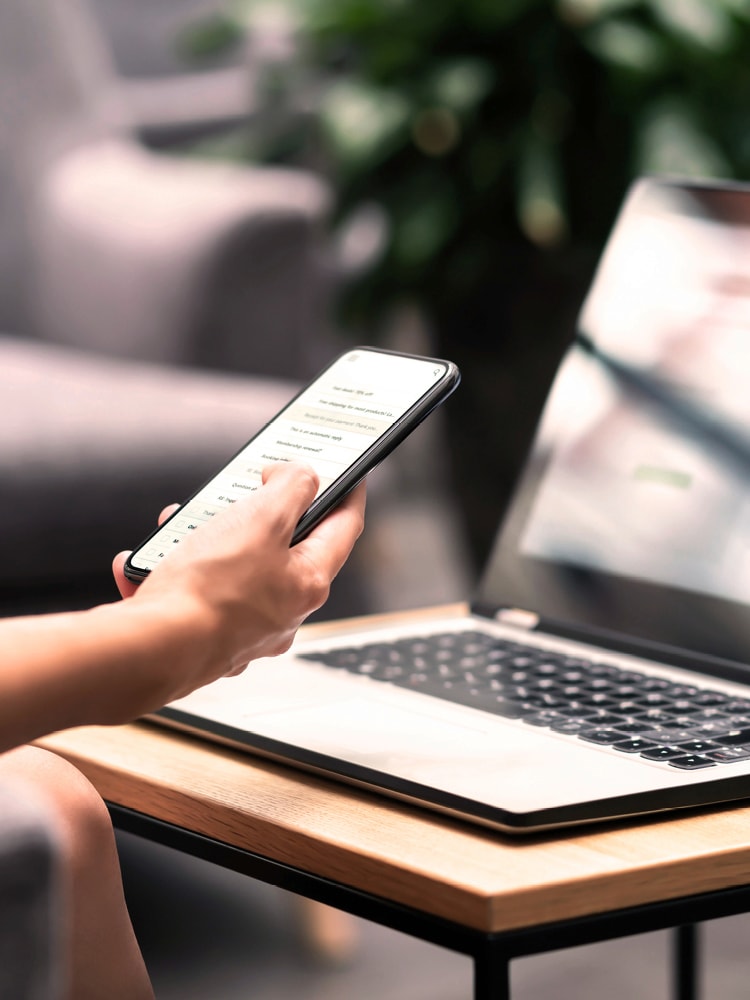 Ein Laptop steht auf einem Couchtisch, davor sitzt eine Frau, die ein Smartphone in der Hand hält