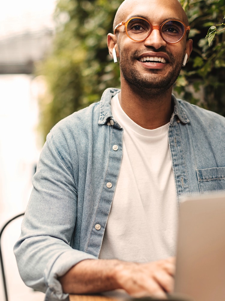 Ein junger Mann mit Brille und Jeanshemd sitzt im freien und arbeitet an seinem Laptop