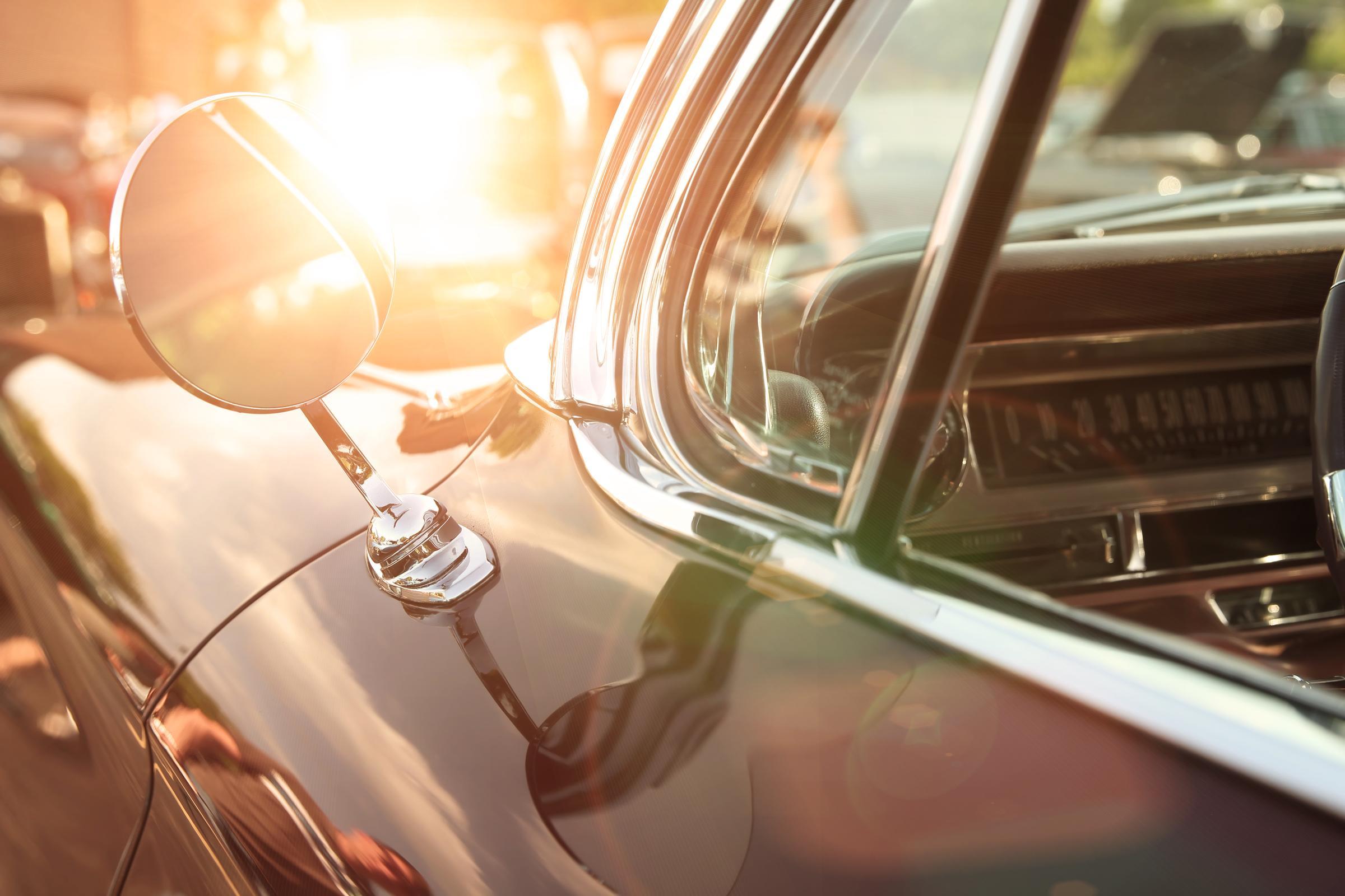 Nahaufnahme eines Seitenspiegels und Fensters eines klassischen Autos, das Sonnenlicht mit einem warmen Glanz reflektiert. Das Bild fängt die glatte, glänzende Oberfläche des Autos und das nostalgische Gefühl von Oldtimern ein.