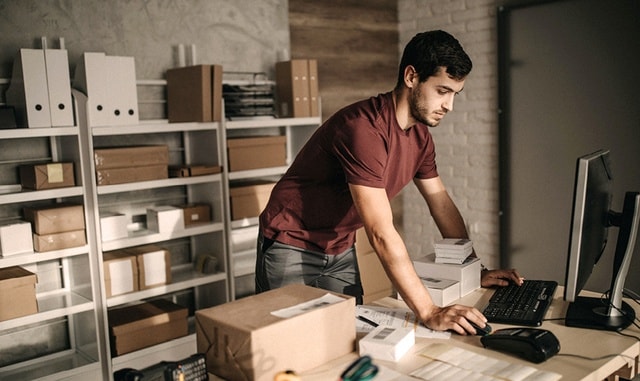 Junger Mann in rotem T-shirt steht an einem Tisch in einem Lager und bearbeitet Bestellungen