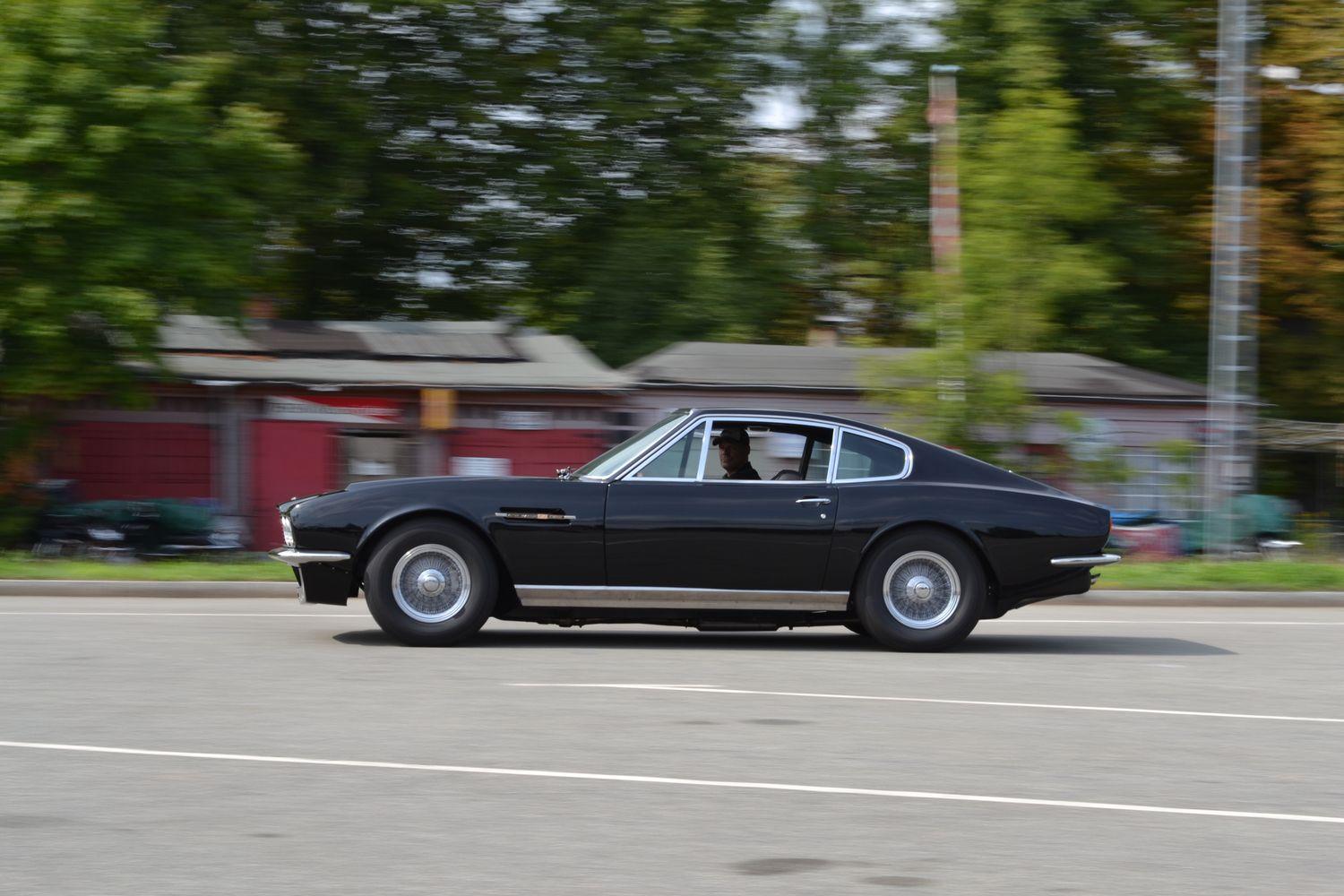 Schwarzer Aston Martin in Bewegung auf einer städtischen Straße mit unscharfem Hintergrund von Bäumen und Gebäuden