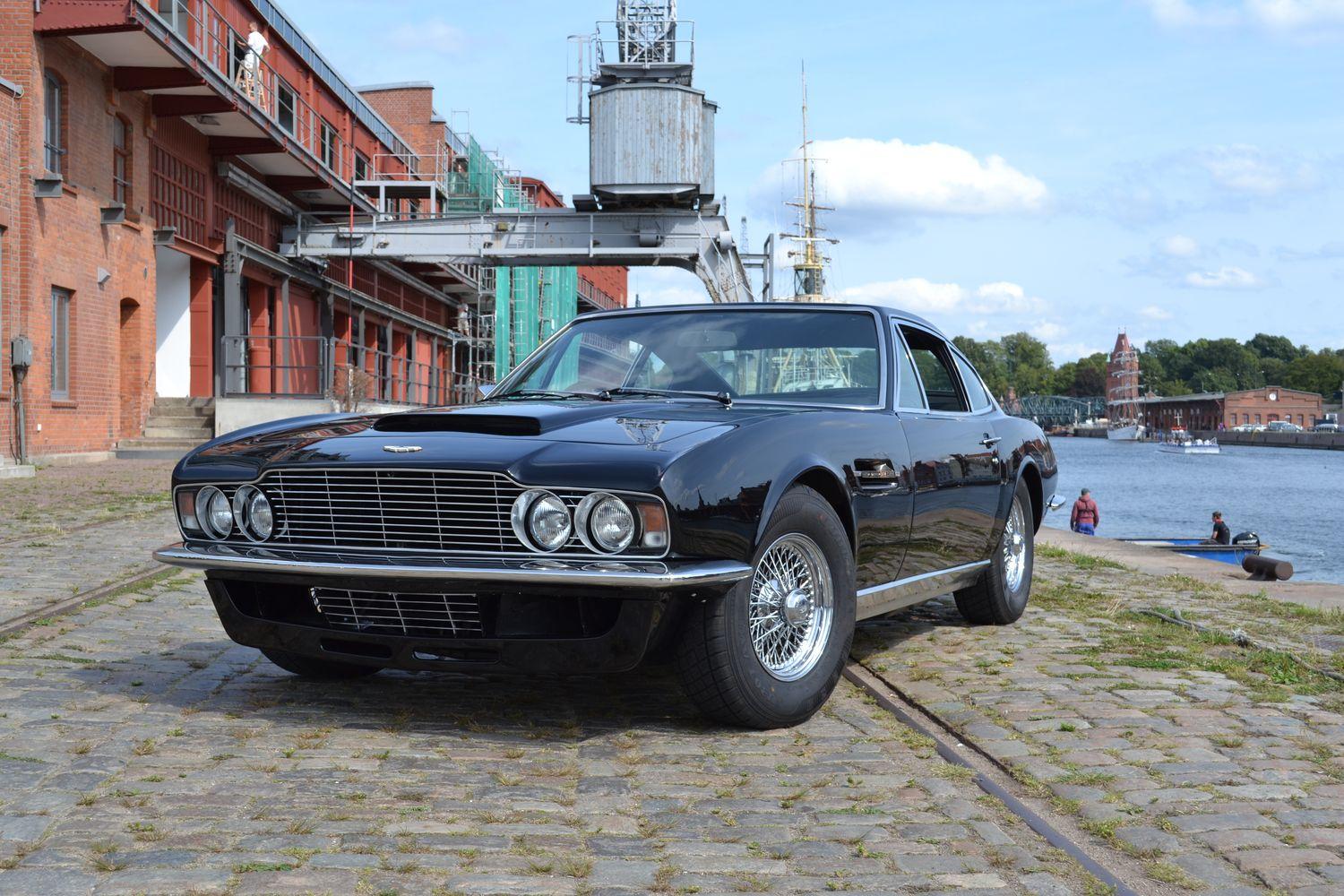 Schwarzer Aston Martin geparkt auf einer gepflasterten Straße vor einem Hafengebäude mit einem Kran im Hintergrund