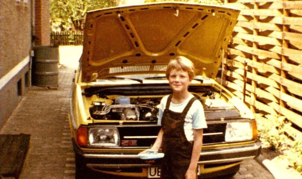 Ein Junge steht neben einem gelben Audi 100 Typ 43 mit geöffneter Motorhaube.