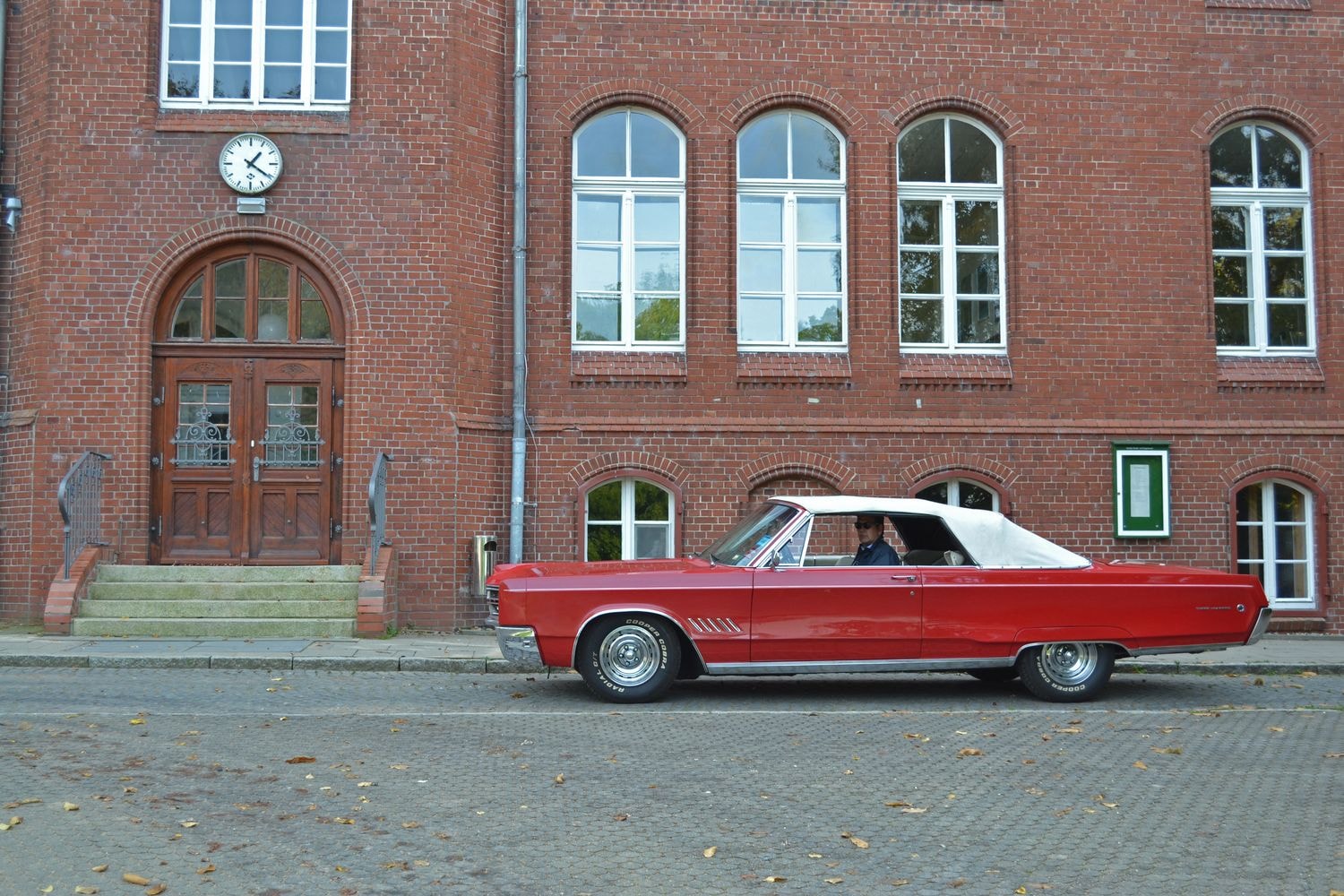 Ein rotes Oldtimer-Cabriolet mit weißem Verdeck steht vor einem historischen Backsteingebäude mit großen Fenstern und einer Uhr über dem Eingang