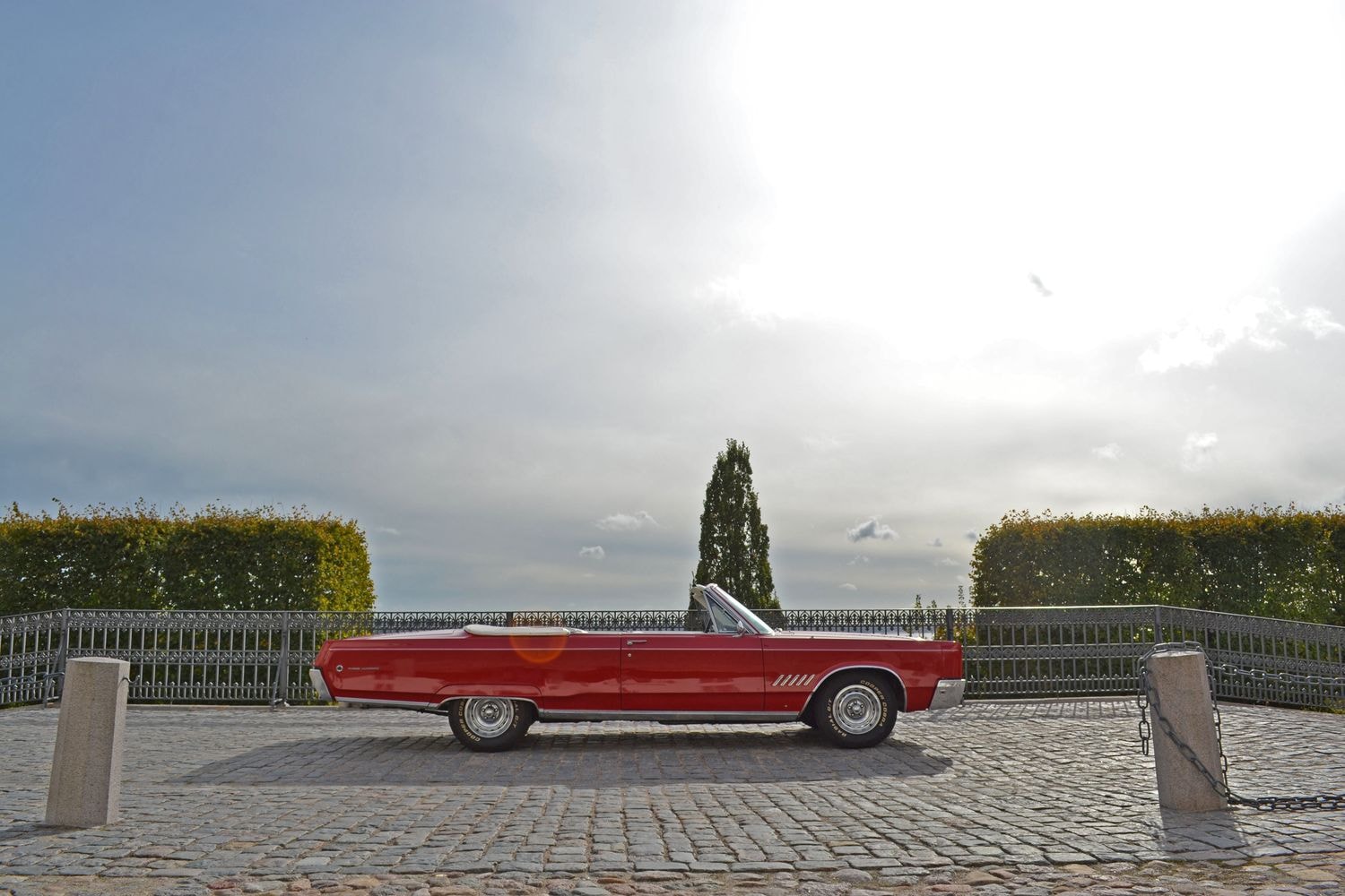 Ein rotes Cabriolet steht auf einem gepflasterten Platz vor einem Zaun und hohen Hecken. Der Himmel ist bewölkt, aber hell