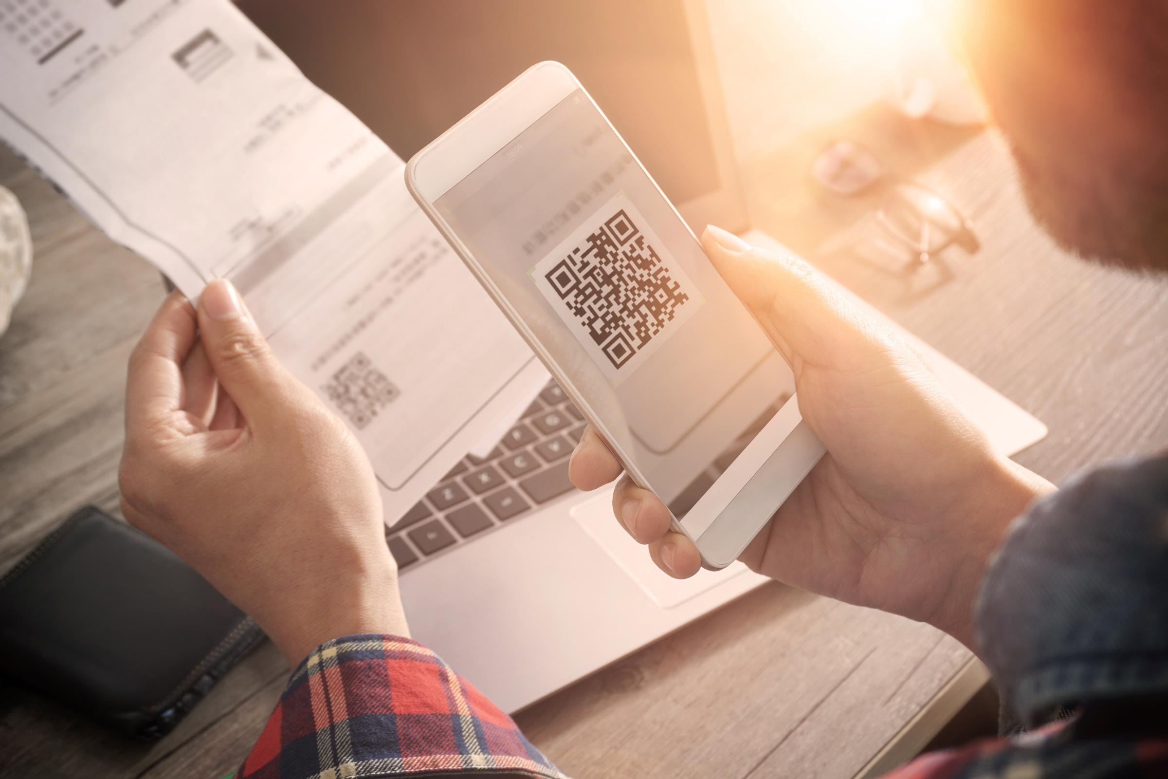 Person scannt mit einem Smartphone einen QR-Code auf einem Papierdokument, im Hintergrund sind ein Laptop und eine Brille sichtbar