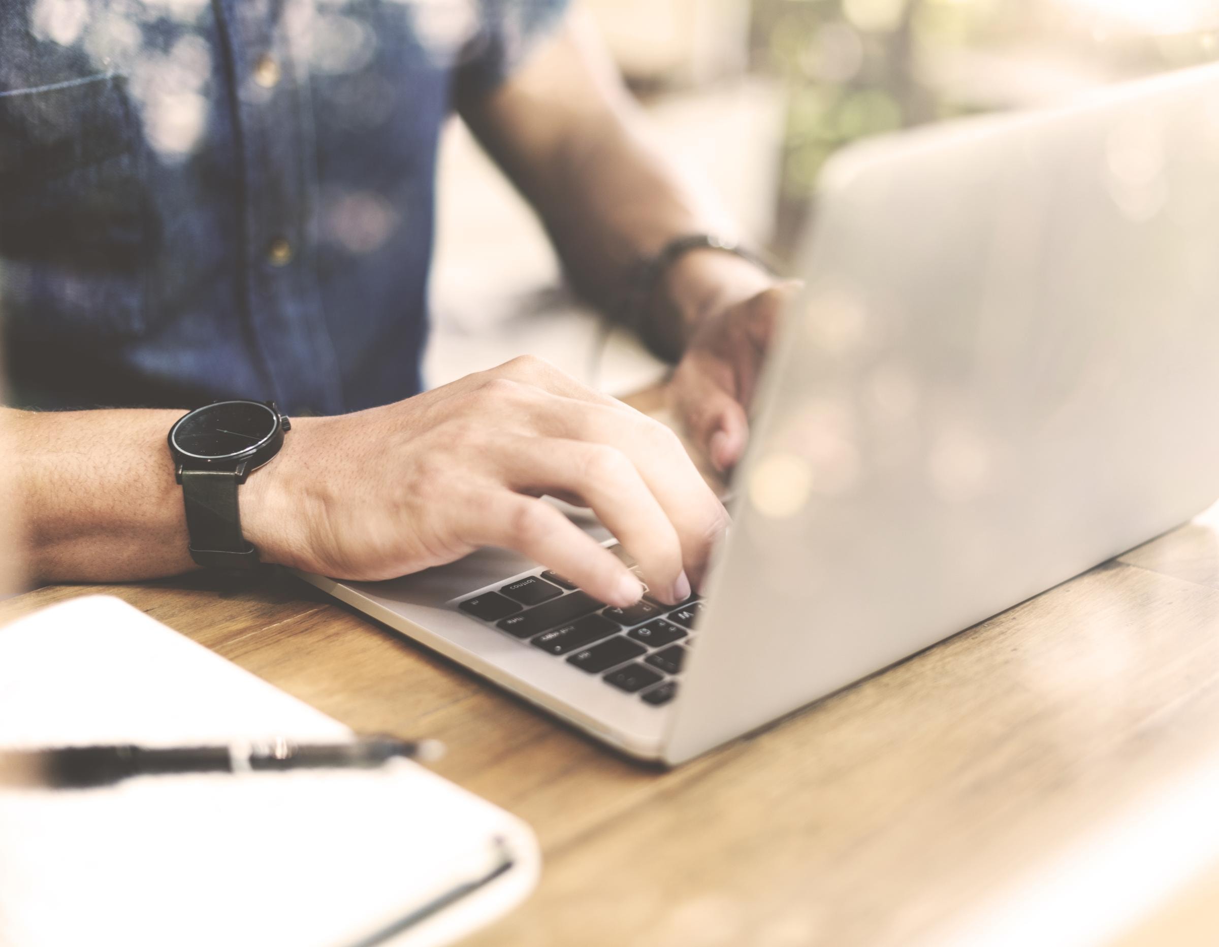 Eine Person, die ein blaues Hemd und eine schwarze Uhr trägt, tipp auf einen Laptop. Das Gesicht der Person ist nicht zu sehen.