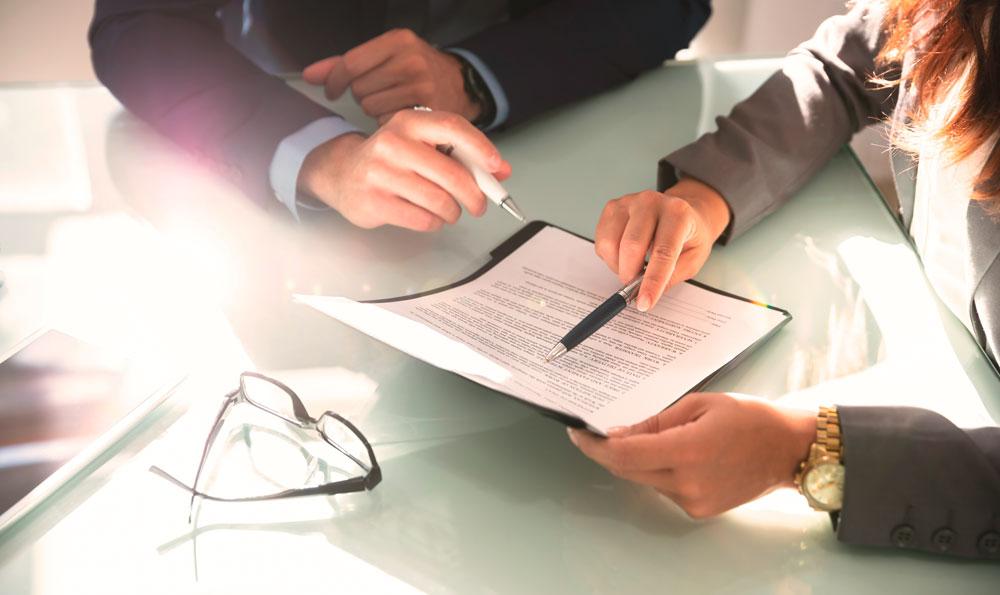 Hände von einer Frau und einem Mann in Business-Kleidung, die an einem Tisch einen Vertrag besprechen; eine Brille liegt daneben.