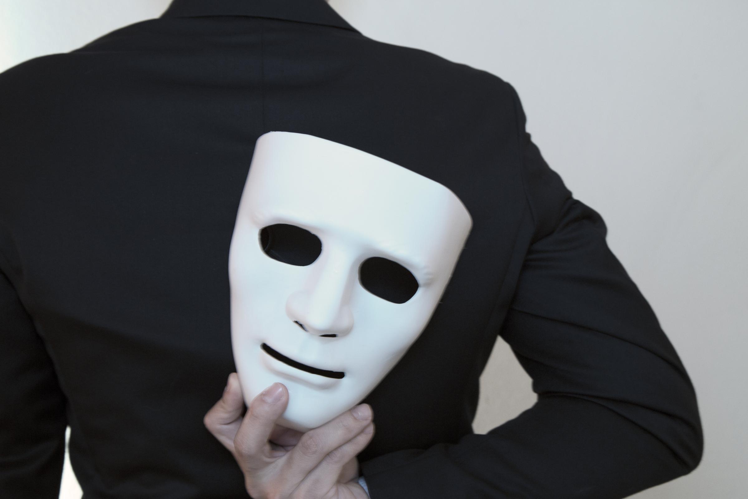 Eine Person in einem schwarzen Anzug hält eine weiße Maske hinter ihrem Rücken. Die Maske ist teilweise verdeckt und die Person ist von hinten zu sehen.