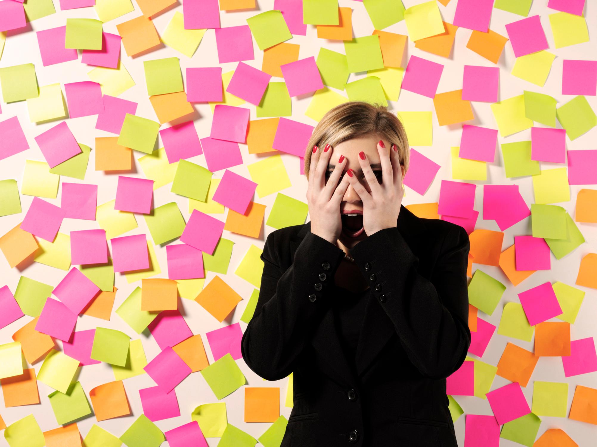 Eine geschockte Frau im Anzug verdeckt ihr Gesicht mit den Händen vor einer Wand voller Post-its.