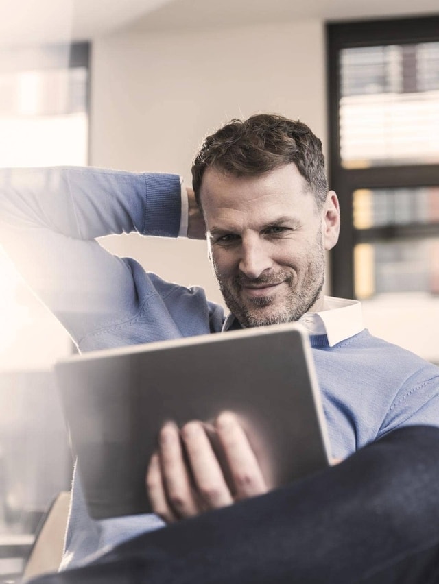 Ein Mann in einem blauen Pullover sitzt entspannt und hält ein Tablet in der Hand. Sein Gesicht ist unscharf gemacht. Im Hintergrund sind Fenster mit Jalousien zu sehen.