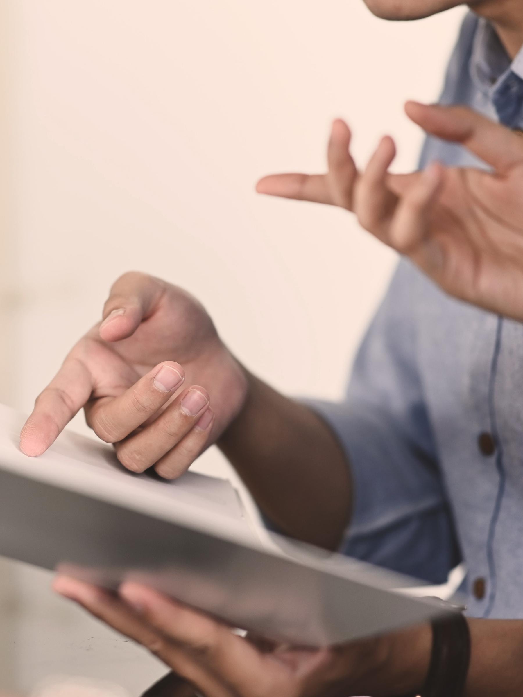 Eine Person trägt ein blaues Hemd und hält ein geöffnetes Buch oder Notizbuch. Eine Hand zeigt auf die Seiten, während die andere Hand gestikuliert.