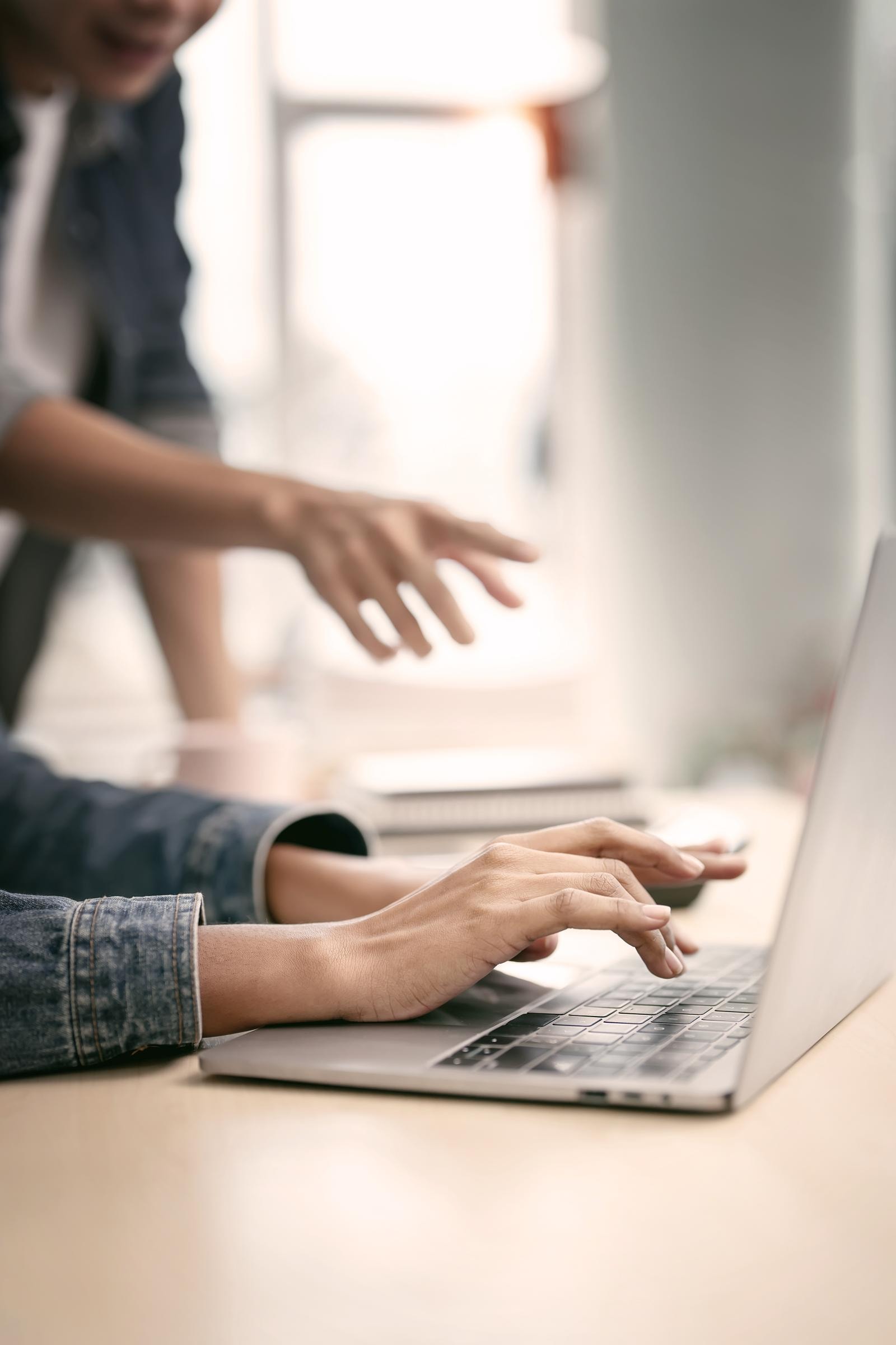 Zwei Personen arbeiten an einen Laptop, eine Person tippt, während die andere im Hintergrund unscharf zu sehen ist.