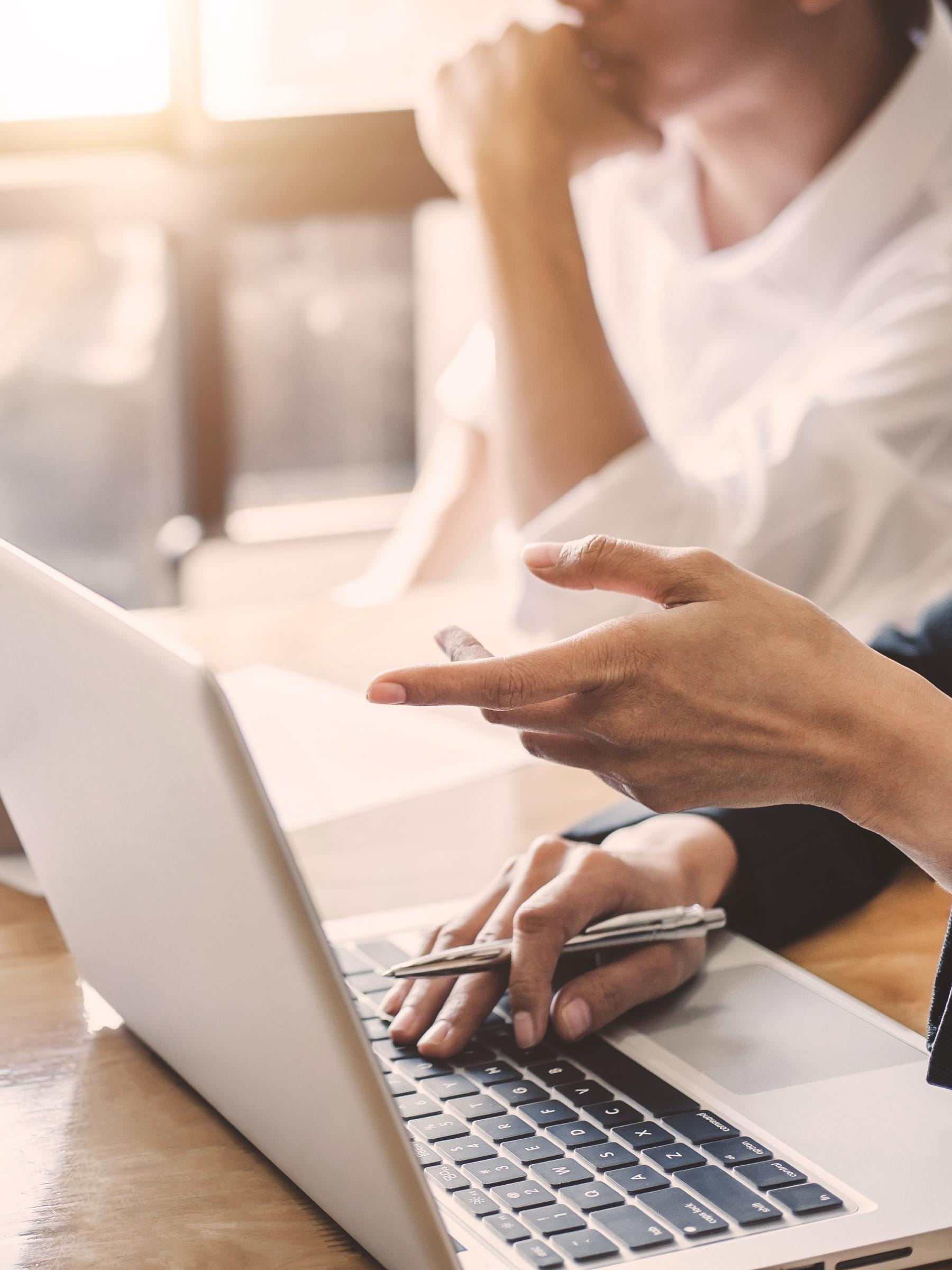 Eine Person arbeitet am Laptop, während eine andere Person, deren Gesicht nicht sichtbar ist, zuschaut.