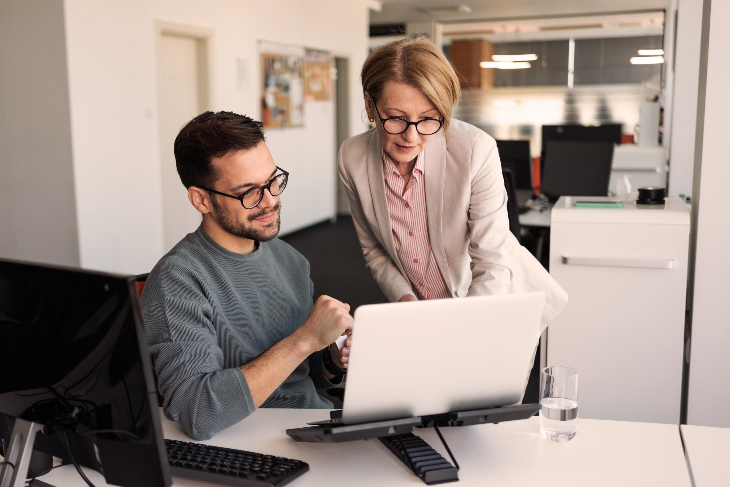 Zwei Personen arbeiten zusammen an einem Computer in einem modernen Büro. Eine Person sitzt und tippt auf der Tastatur, während die andere Person steht und auf den Bildschirm schaut.